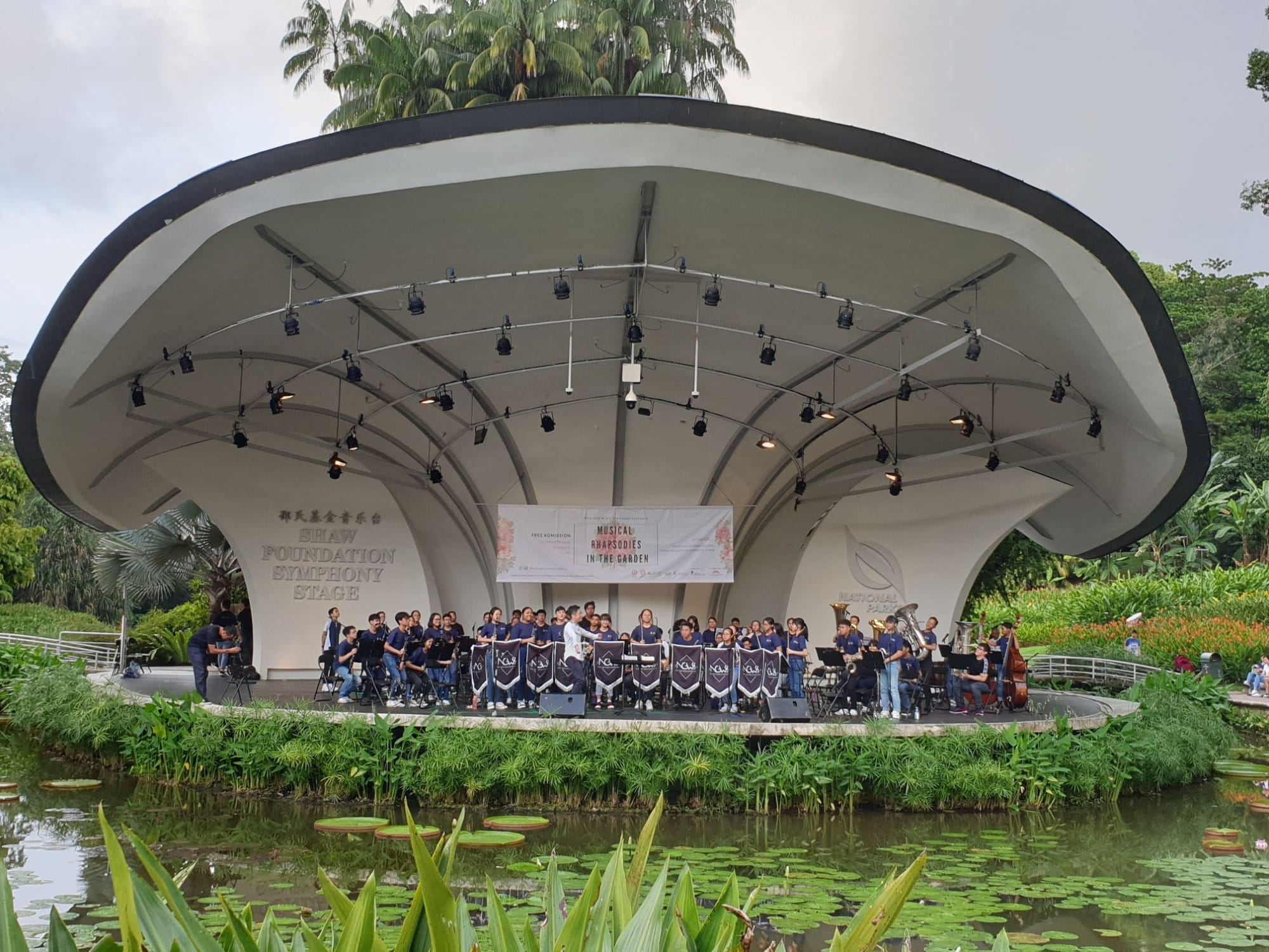 A band performs on the Shaw Foundation Symphony Stage in a garden, surrounded by a pond with lily pads and lush greenery.